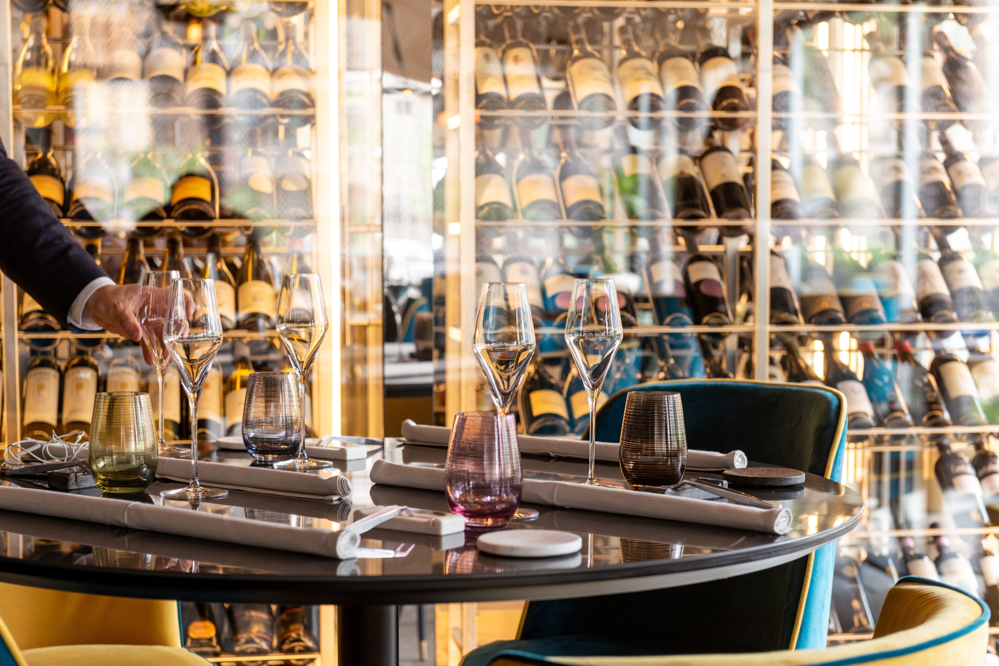 A restaurant table set with wine glasses and bottles, with a person serving.