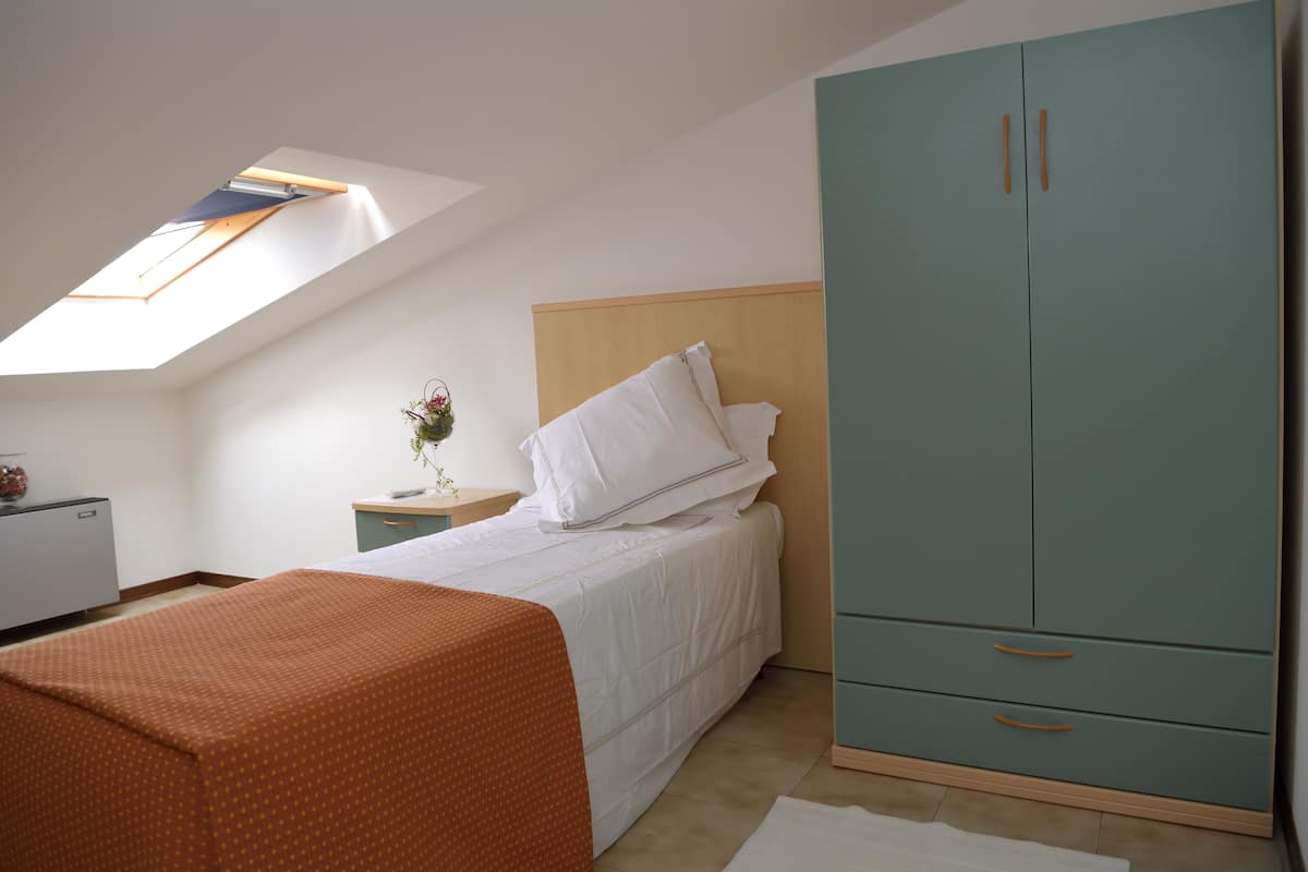 Une chambre avec un puits de lumière, un lit avec une literie blanche, une armoire turquoise et une petite table de chevet sur laquelle se trouve un vase.