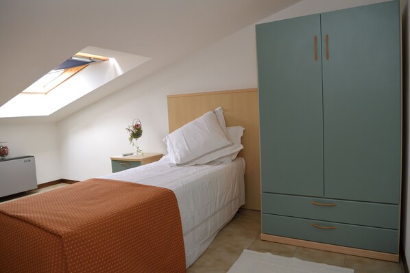 A bedroom with a skylight, a bed with white bedding, a teal wardrobe, and a small bedside table with a vase.