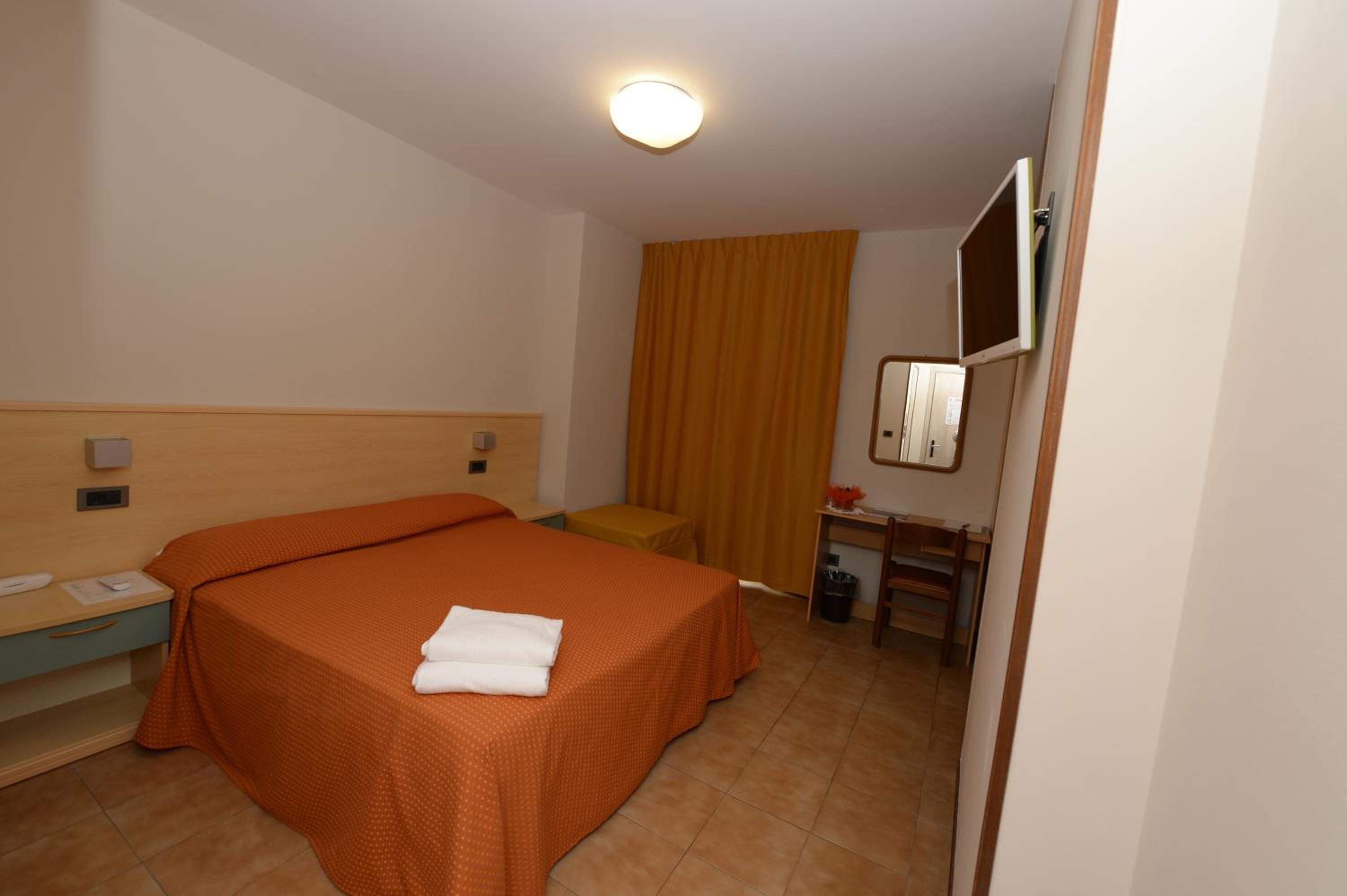 Une chambre d’hôtel avec un lit simple, un petit bureau avec une chaise, un miroir et un téléviseur fixé au mur.