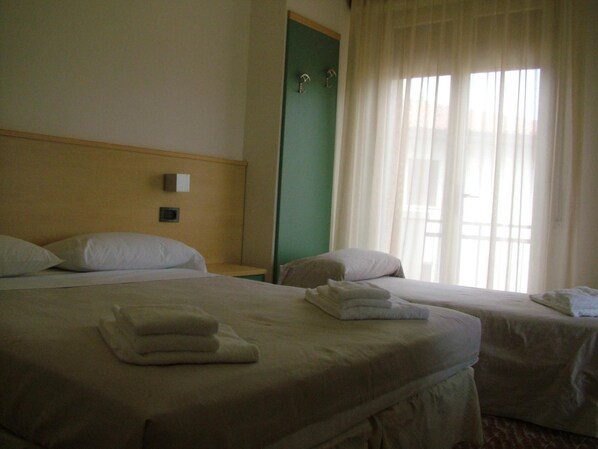 Ein Hotelzimmer mit zwei Betten, einem Nachttisch, einem an der Wand montierten Telefon und einem Fenster mit durchsichtigen Vorhängen.