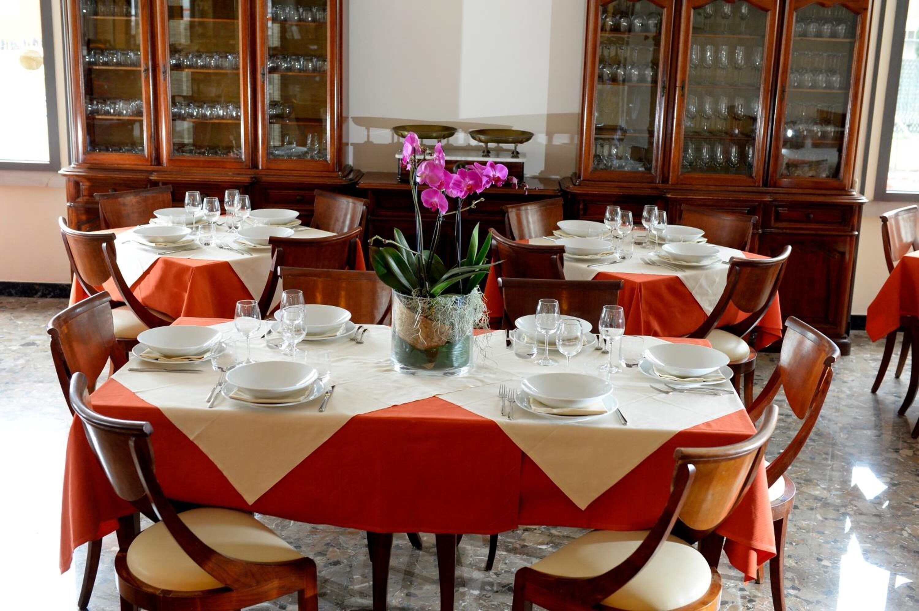 Ein Essbereich mit runden Tischen, gedeckt für sechs Personen. Jeder Tisch ist mit einer weißen Tischdecke und orangefarbenen Servietten versehen. Holzstühle umgeben die Tische, und in der Mitte steht eine große Vase mit violetten Blumen.