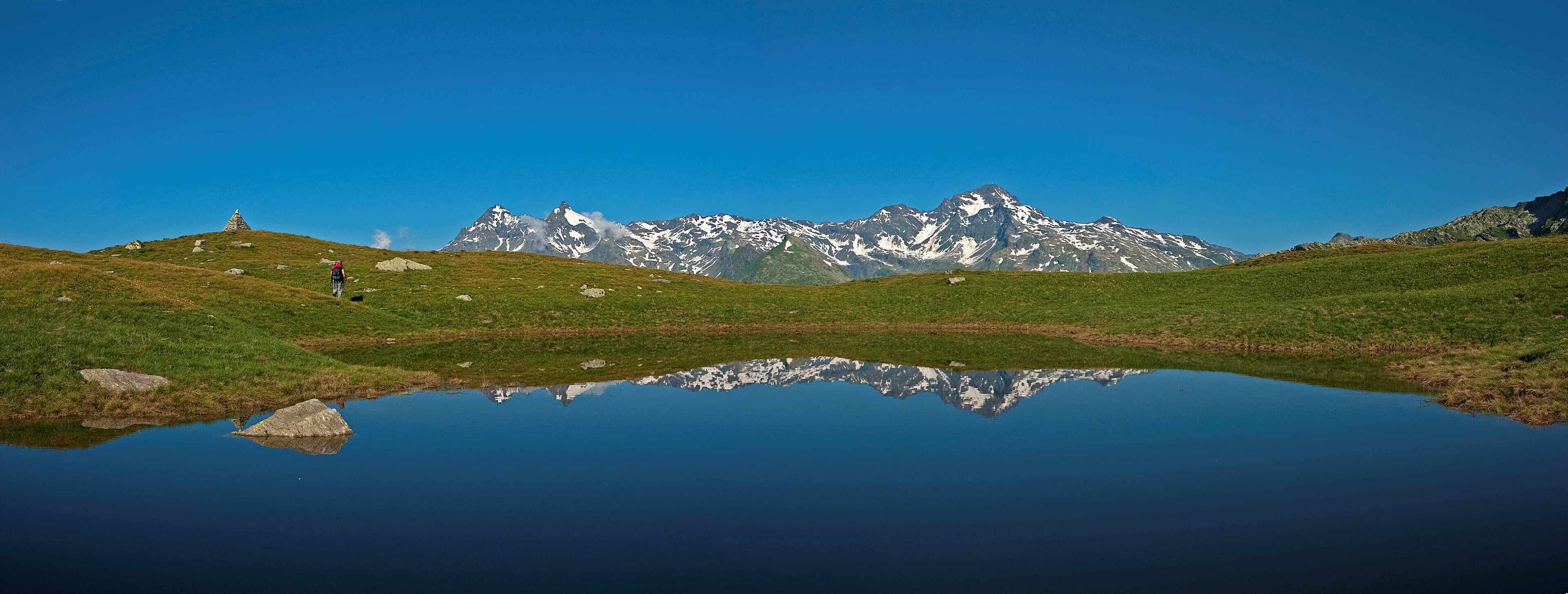 A mountain landscape with a clear reflection in a calm body of water, a person hiking, and a small structure on a hill.