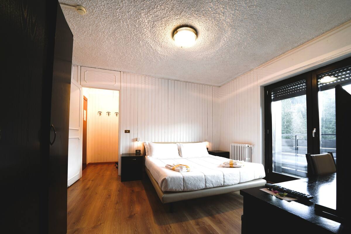 A bedroom with a wooden floor, a bed with white bedding, a nightstand, a desk with a keyboard, and a window with a view of greenery.