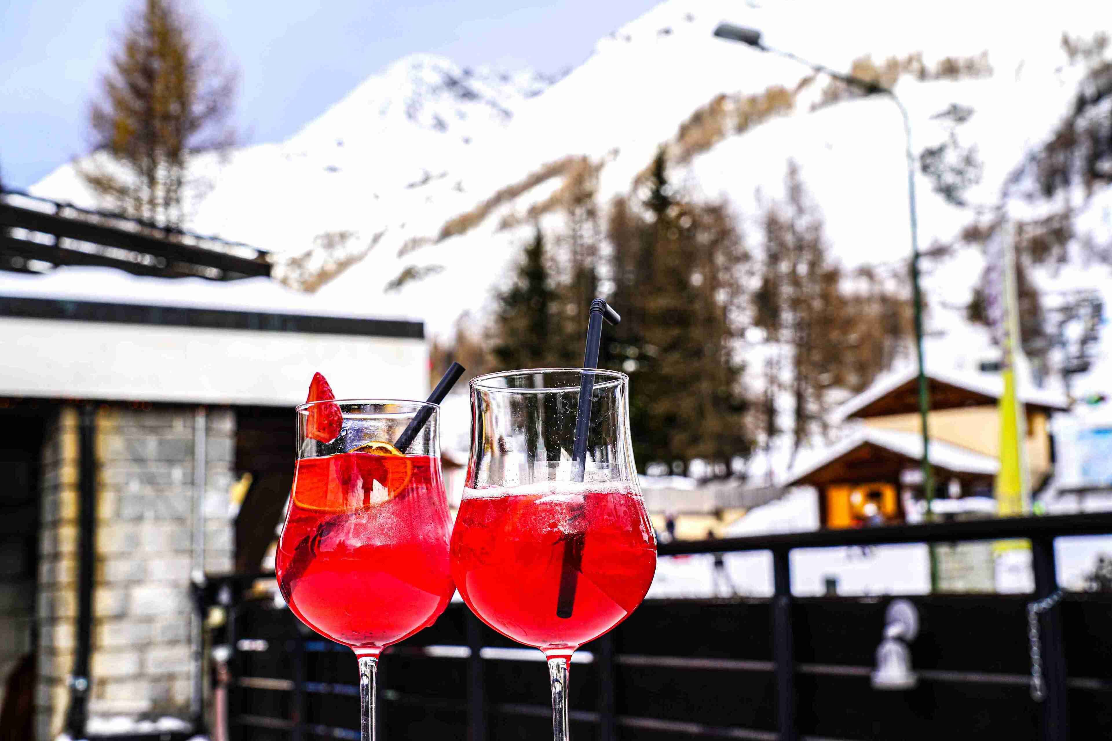 Two glasses of red beverage with straws and a slice of orange, set against a snowy mountain backdrop.