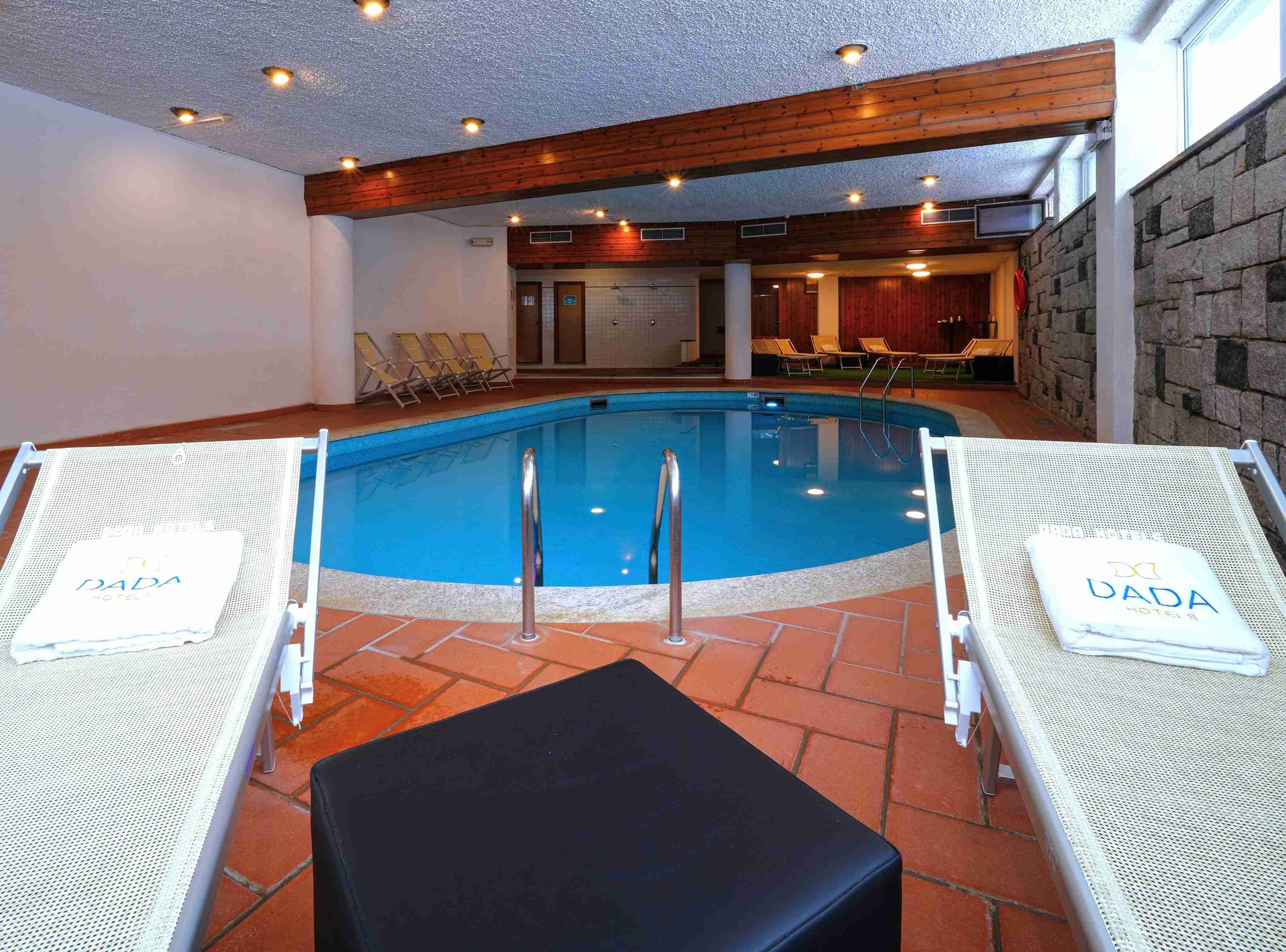 An indoor swimming pool with two lounge chairs, a bench, and a wooden ceiling.