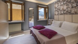 A modern bedroom with a large bed, two windows, a radiator, and a cityscape view. - Hotel Aurora (Malcesine)