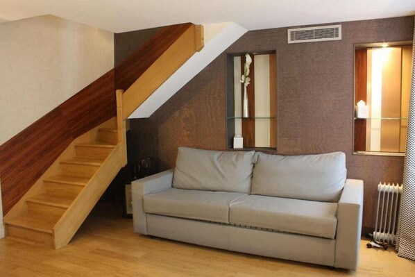 A living room with a wooden staircase, a beige sofa, a mirror, and a radiator. - Hôtel Le Malown (Paris)
