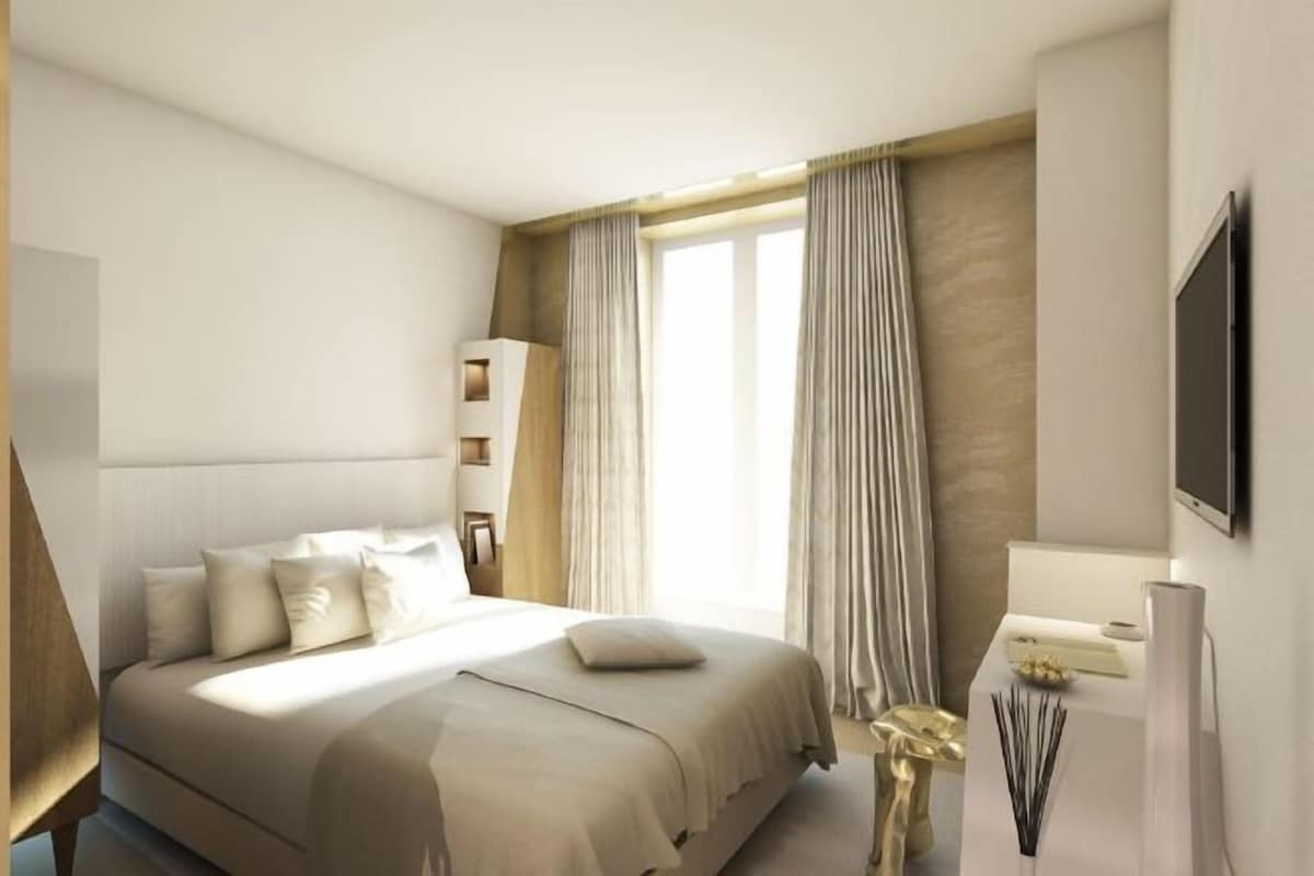 A modern hotel room with a large bed, a bedside table with a lamp, a TV mounted on the wall, and a window with curtains.