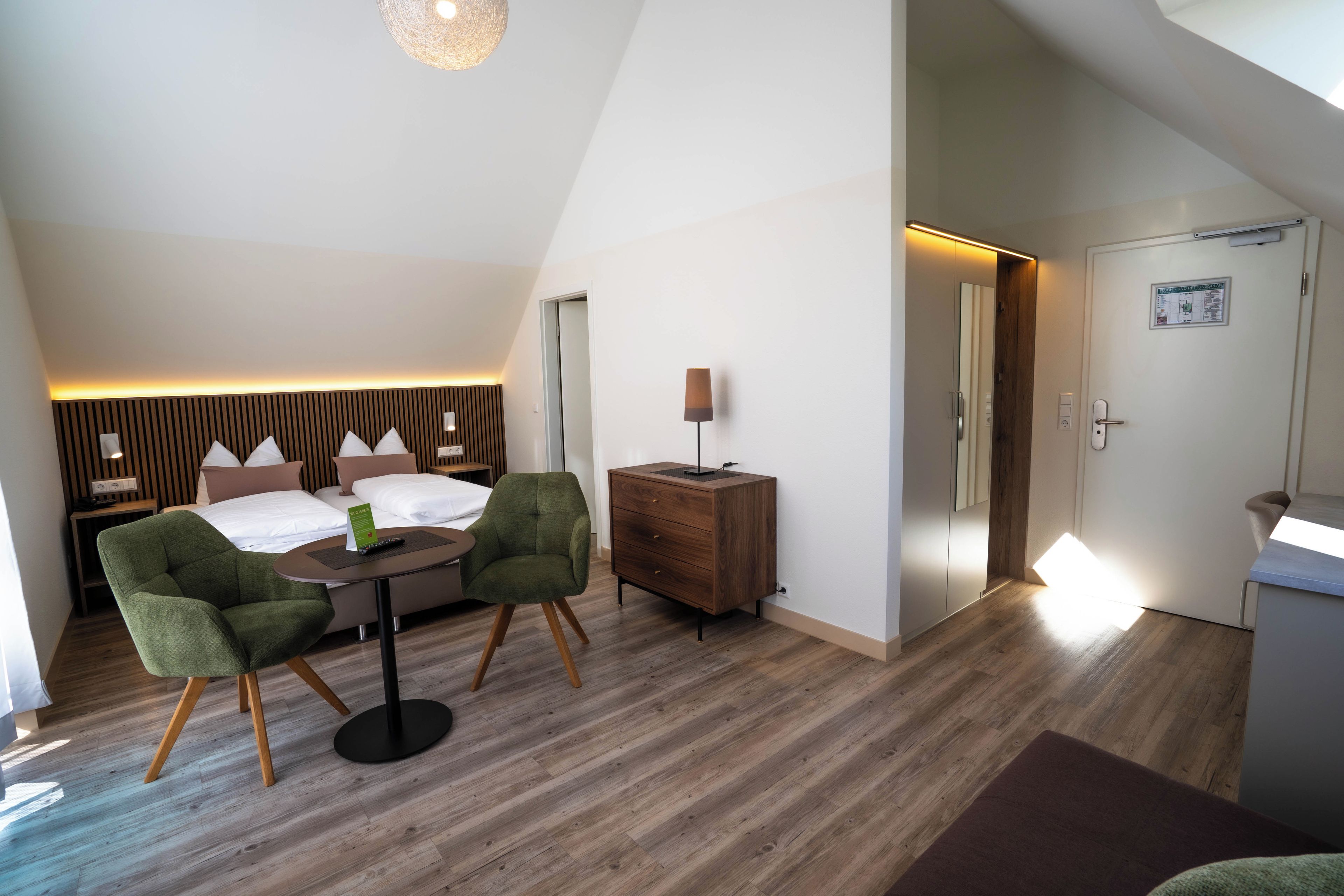Una habitación de hotel moderna con una cama, dos sillones verdes, una mesita, un aparador de madera y un escritorio con una lámpara.