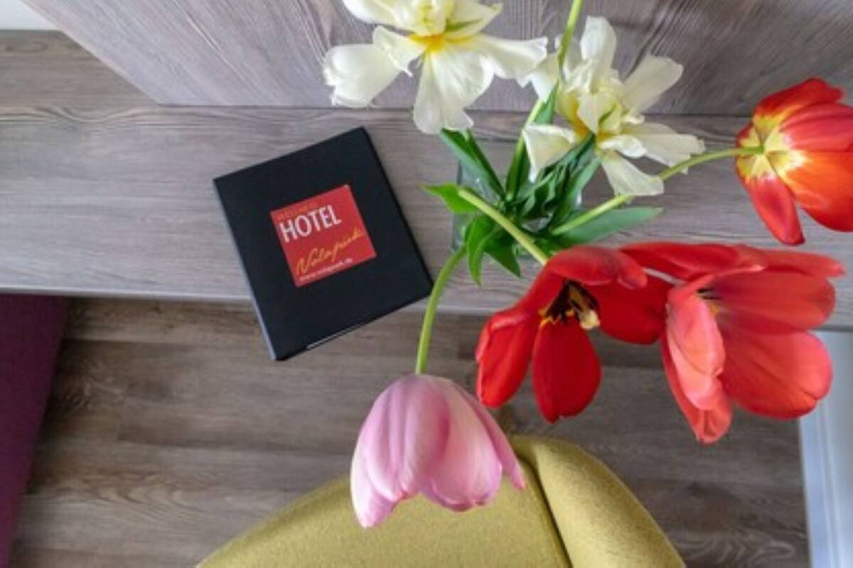 Habitación de hotel con piso de madera, una caja negra con la etiqueta "HOTEL" y un ramo de flores que incluye tulipanes rojos y lirios blancos.