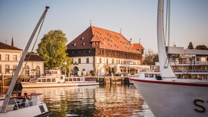 A historic building with a red roof and a white facade, docked boats, and a calm water body. - Hotel Volapük (Konstanz)