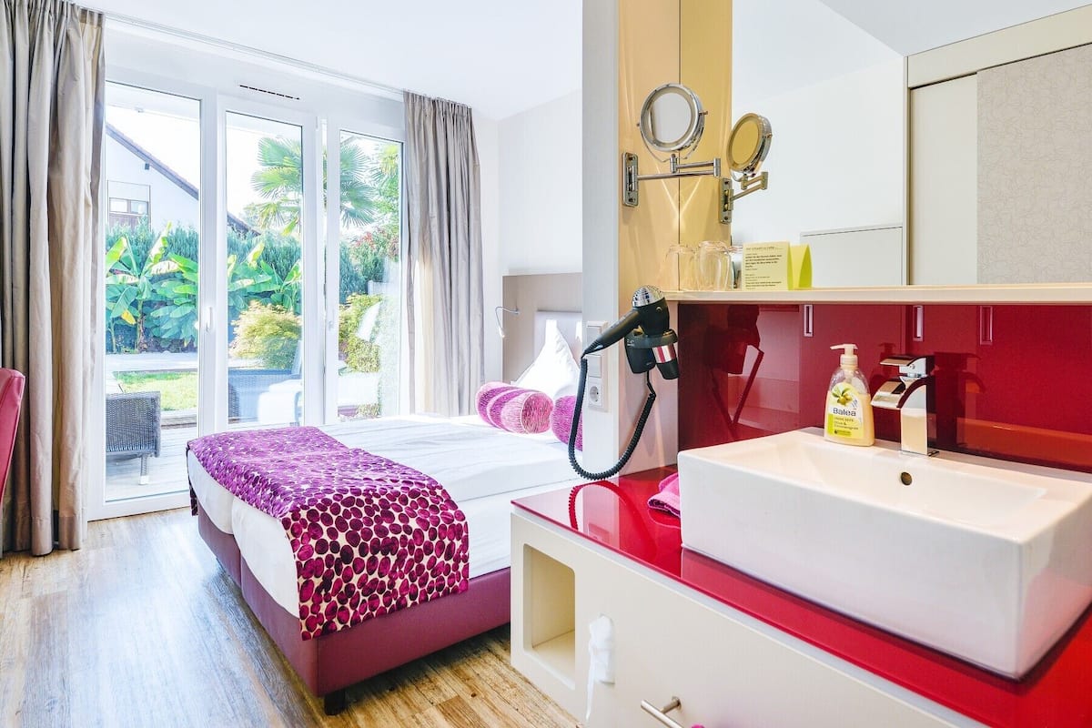 Habitación de hotel con cama, una silla roja, baño con lavamanos y espejo, y vista a zonas verdes.