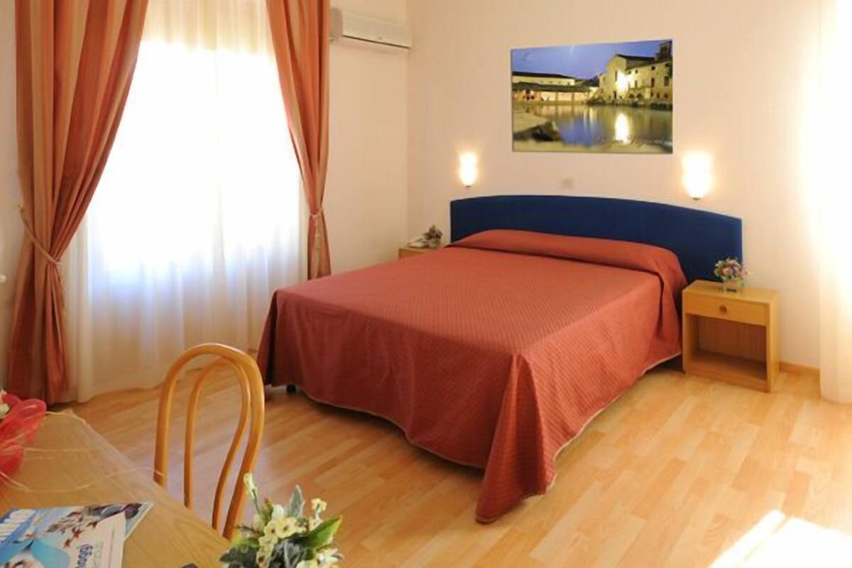 A hotel room with a bed, a desk, and a chair. There is a window with curtains, a picture on the wall, and a nightstand with a lamp.