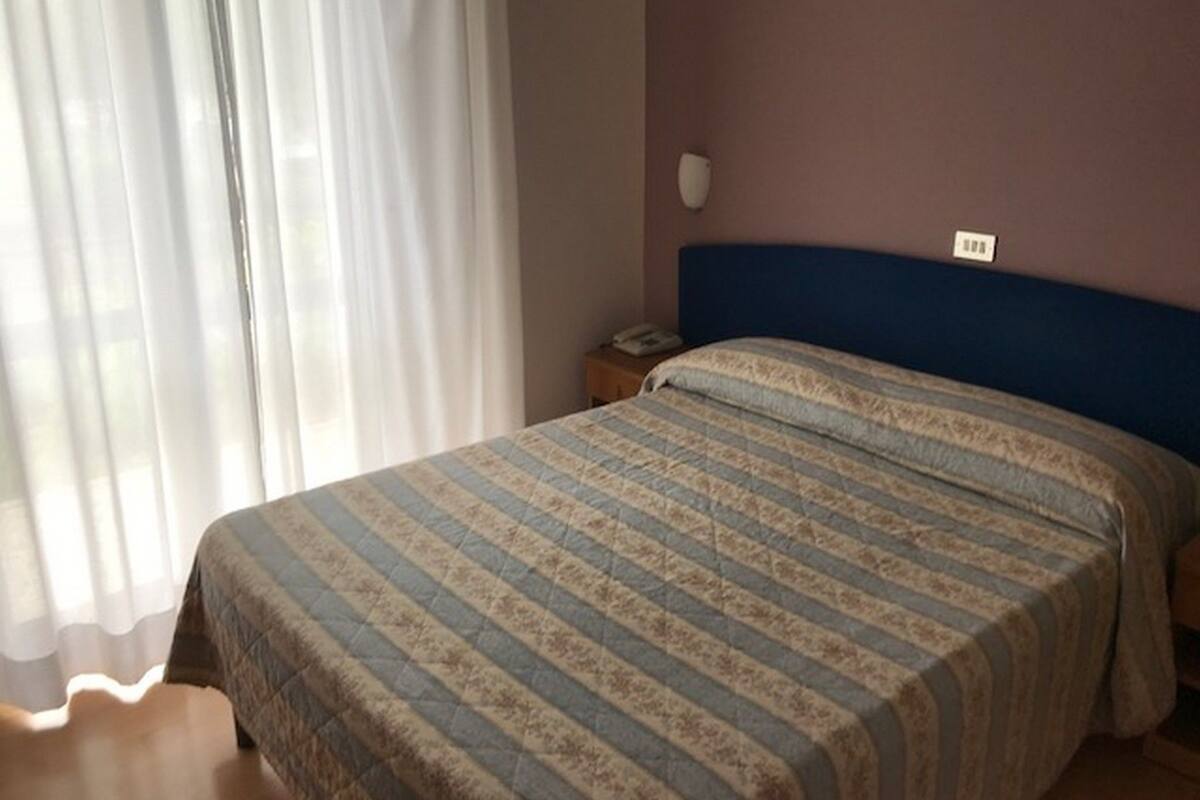 A bedroom with a bed, a nightstand, a wall socket, and a window with curtains.