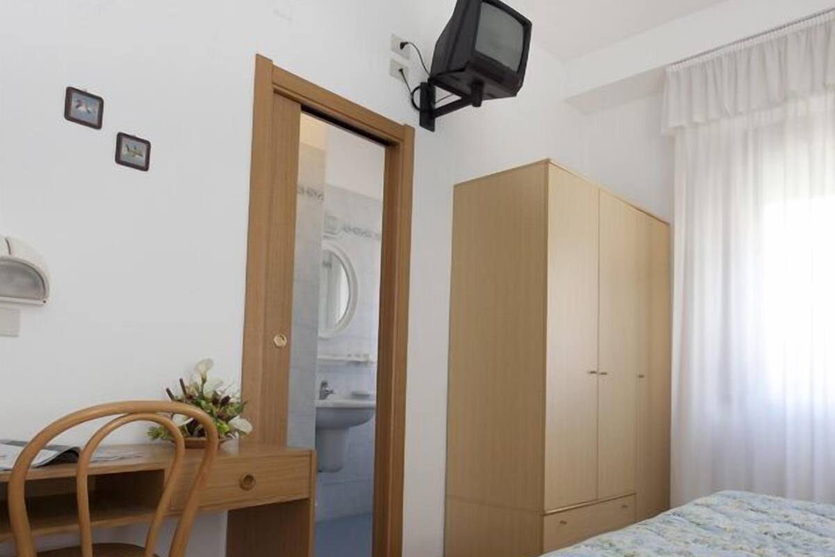 A bedroom with a bed, a wooden wardrobe, a desk with a chair, and a television mounted on the wall.