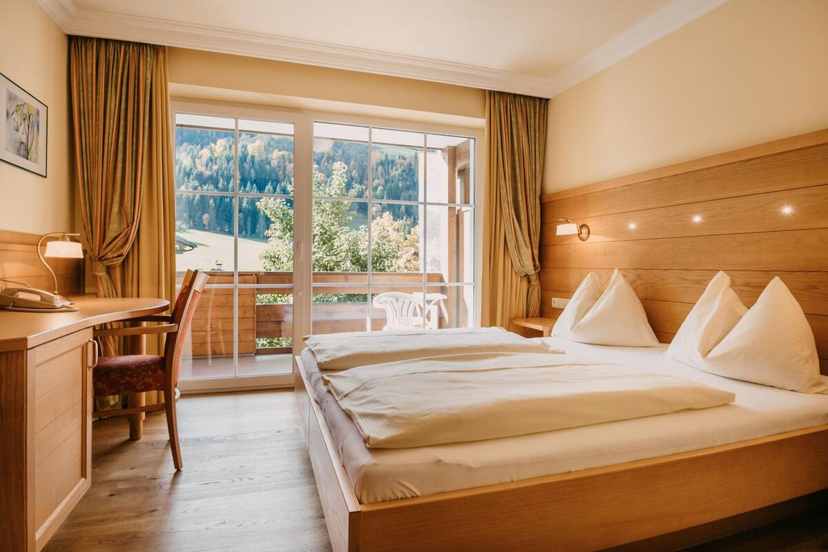 Habitación de hotel con cama de madera, un escritorio y una silla. Hay un ventanal con cortinas, y se ve un paisaje de árboles y montañas.