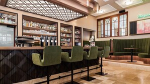 Zona de bar con taburetes tapizados en verde, estantes llenos de botellas y vasos, y un piano.