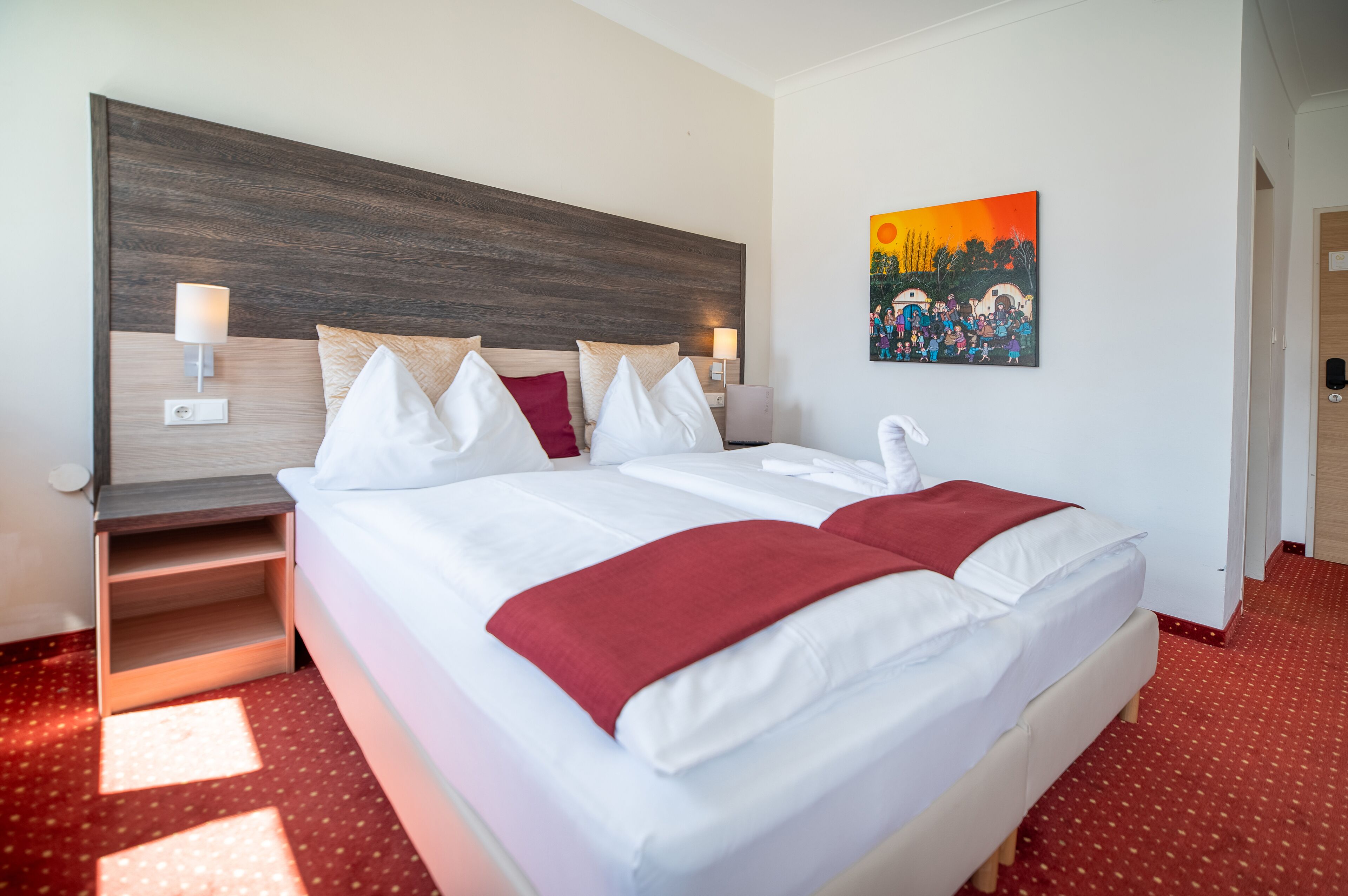 Quarto de hotel com uma cama grande, cabeceira de madeira, tapete vermelho e um quadro na parede.