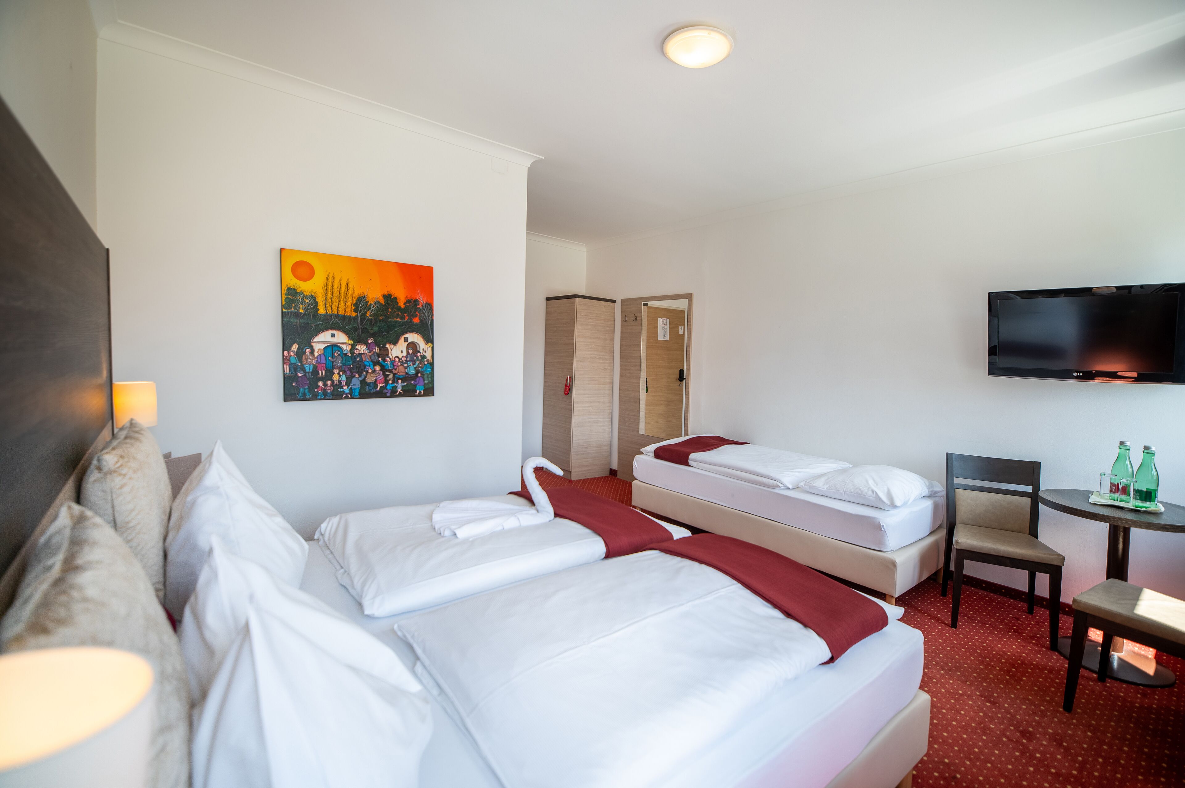 A hotel room with two beds, a TV, a small table with bottles, and a painting on the wall.