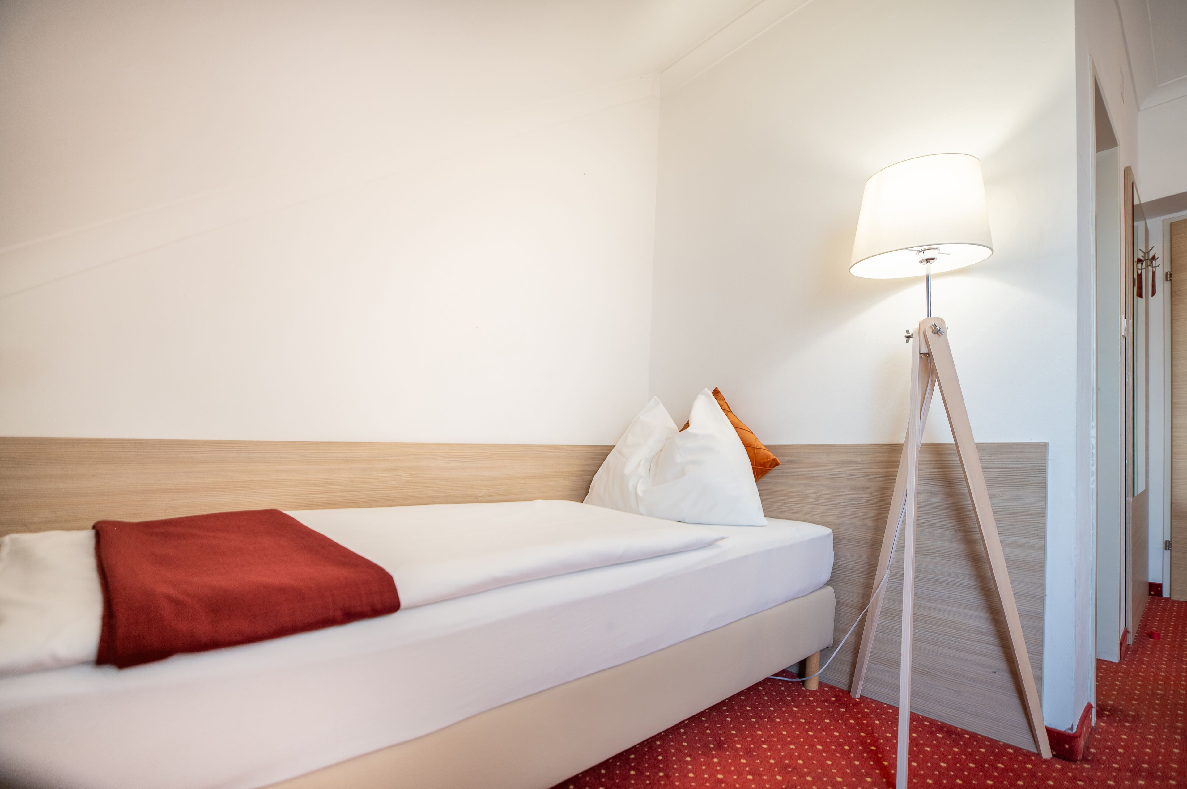 A hotel room with a single bed, a bedside lamp, and a red carpet.
