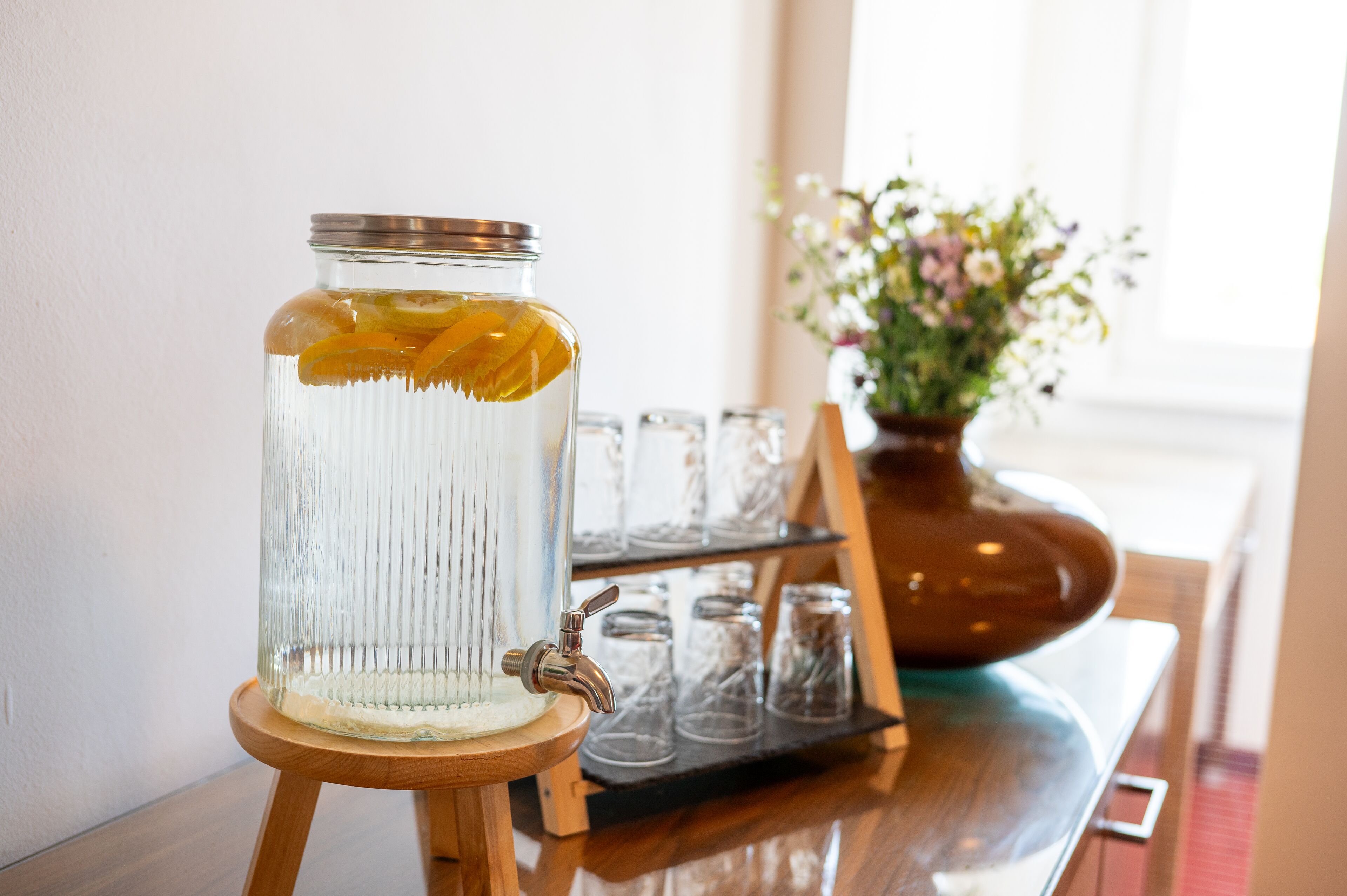 Un frasco de vidrio transparente con tapa metálica, lleno de líquido y rodajas de naranja, sobre un soporte de madera.