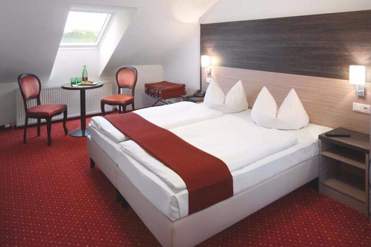 A hotel room with a large bed, a desk, a chair, and a small table with a bottle and glasses.