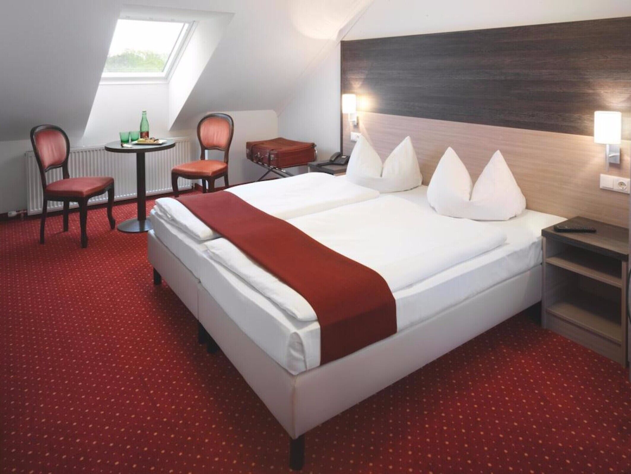 A hotel room with a large bed, a desk, a chair, a small table, and a view of the outdoors through a skylight.