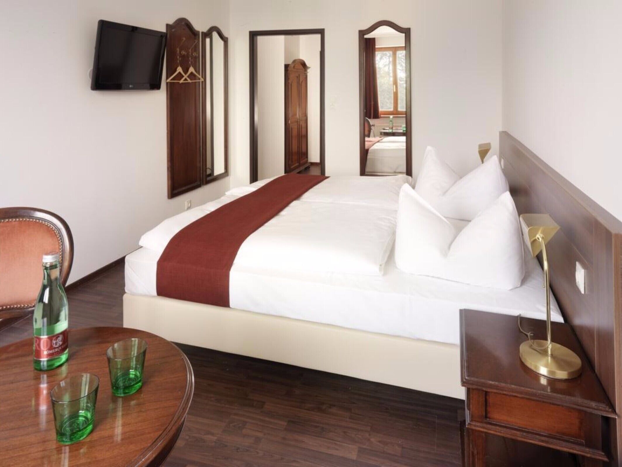 A hotel room with a large bed, a wooden nightstand, a chair, and a table with glasses.