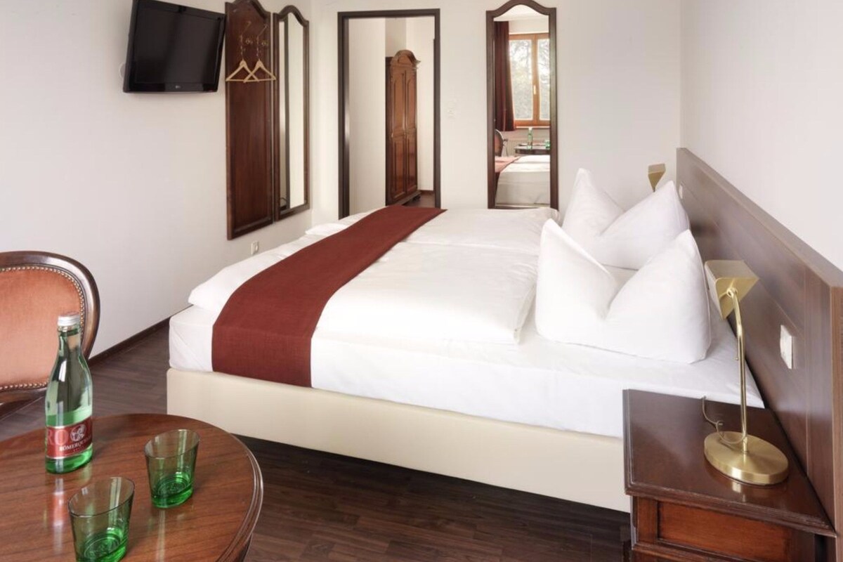 A hotel room with a large bed, a wooden nightstand, a chair, a table with glasses, and a TV on the wall.