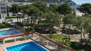 Una piscina rectangular con un borde de baldosas y una barandilla metálica, rodeada de árboles y con vistas a una playa y edificios al fondo.