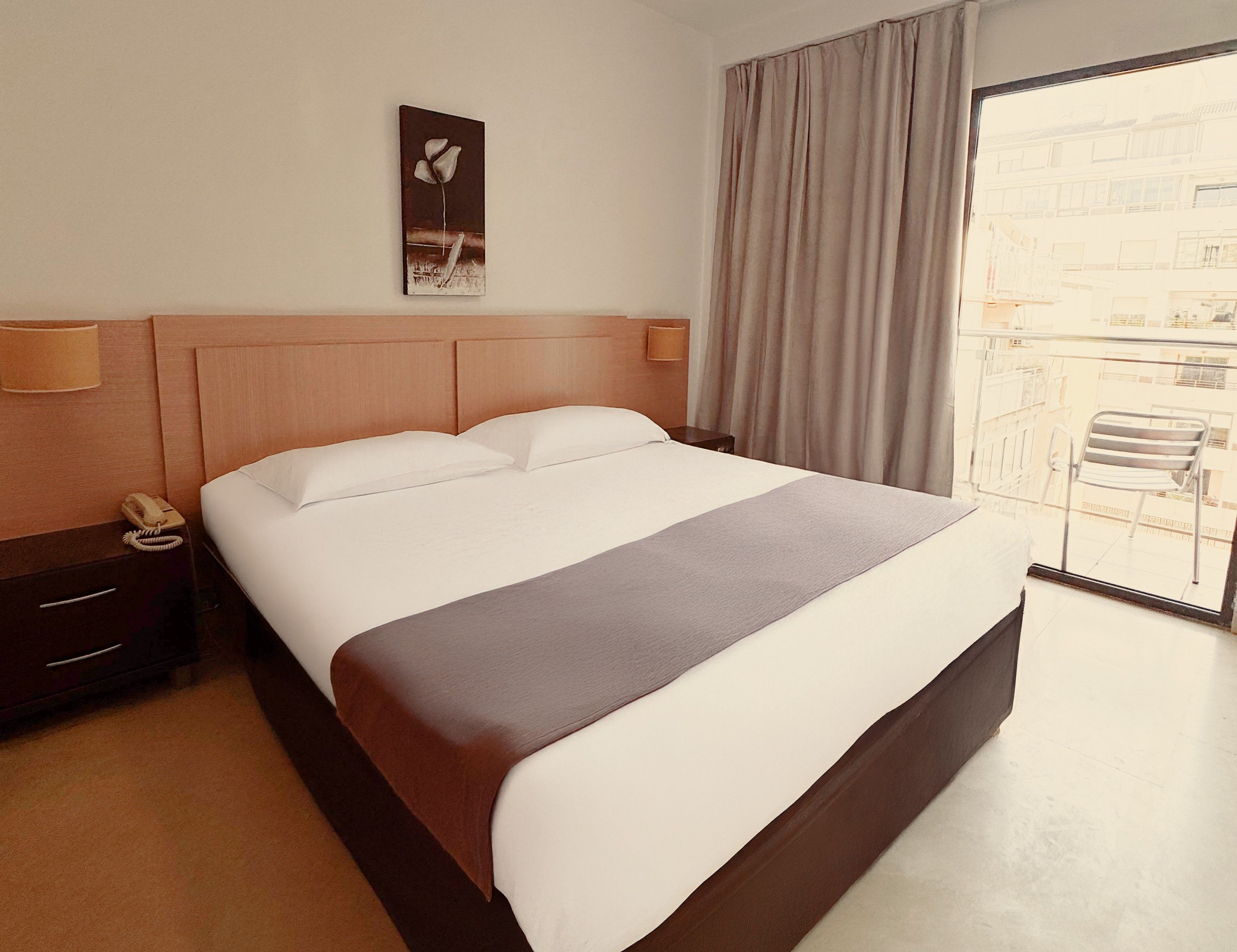 A hotel room with a bed, a nightstand with a phone, a painting on the wall, and a view through the window.