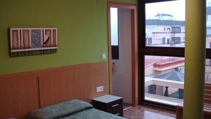 A room with a bed, a small bedside table, a wall-mounted shelf, and a window with a view of buildings. - Primavera (Benidorm)