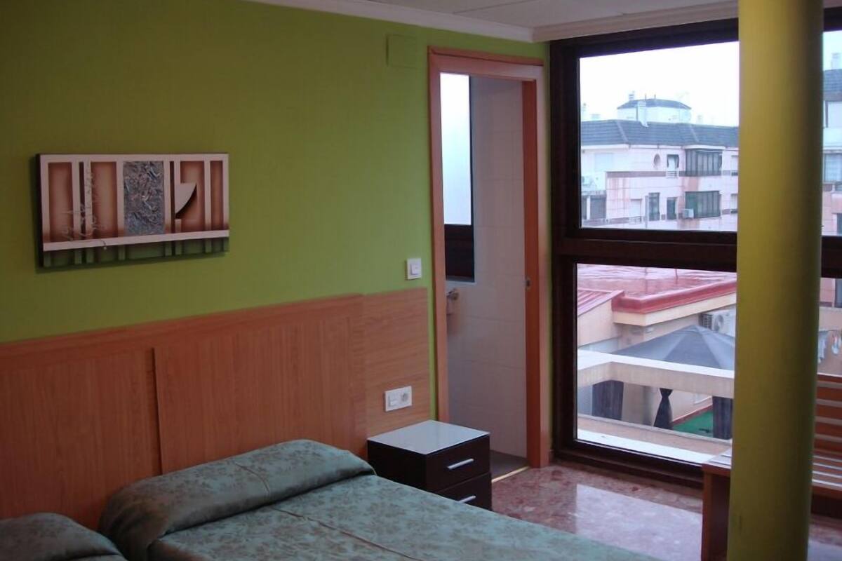 Quarto com cama, mesa de cabeceira pequena, prateleira na parede e janela com vista para edifícios.