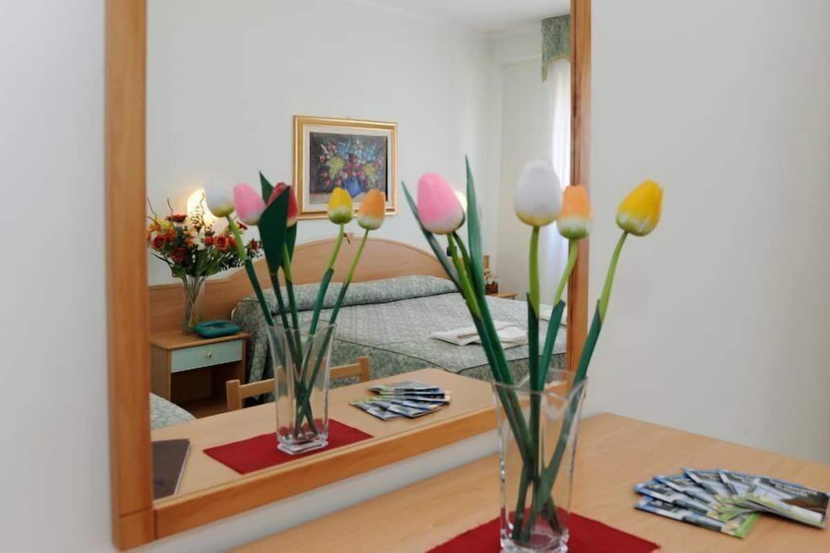 A bedroom with a bed, a bedside table with a lamp, a mirror, and a vase with flowers.