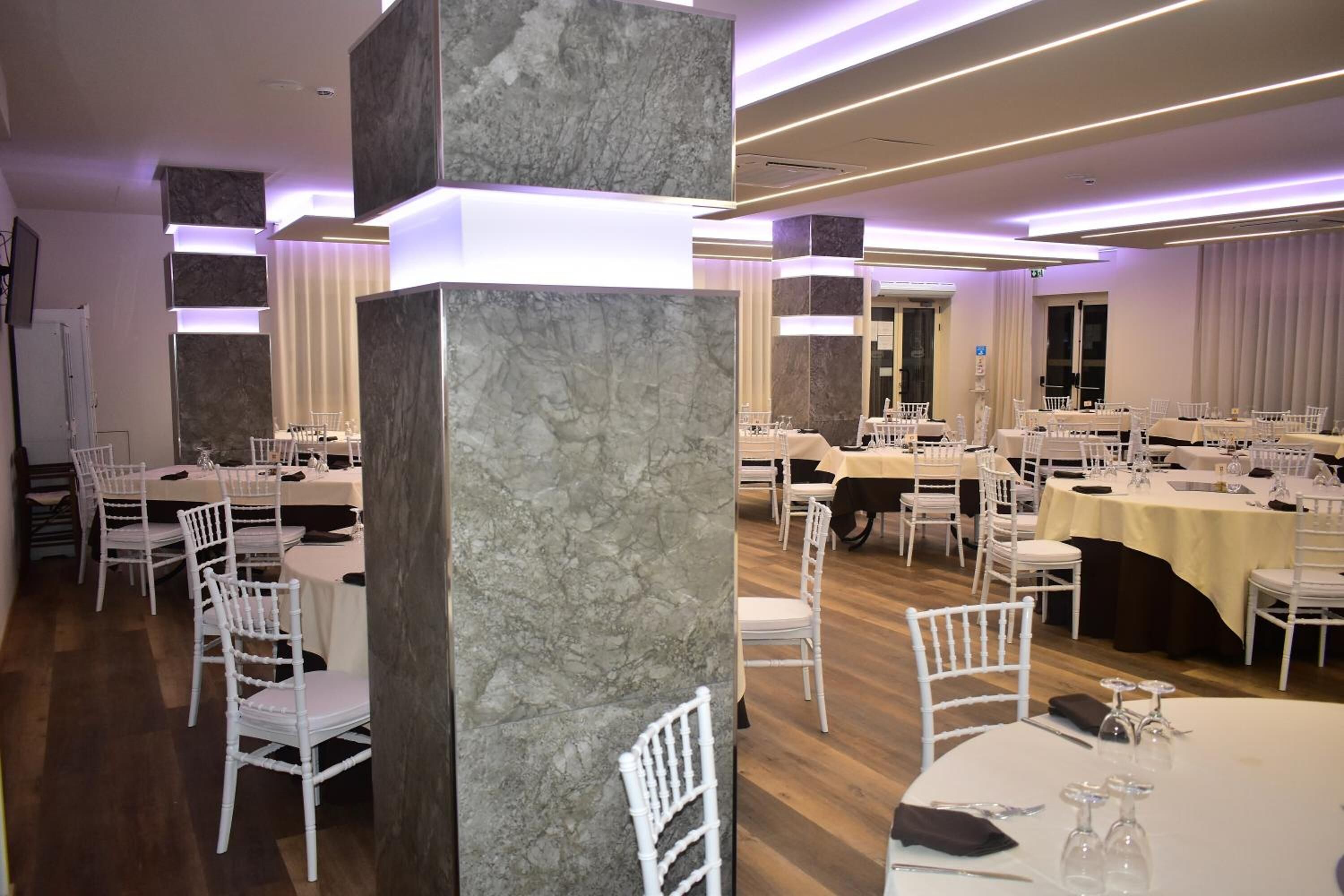 Un comedor amplio con varias mesas y sillas blancas, pilares de mármol e iluminación moderna.