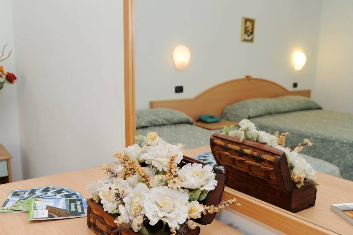 A bedroom with a wooden headboard, a bed with green bedding, a bedside table with a vase of flowers, and a mirror reflecting the room.
