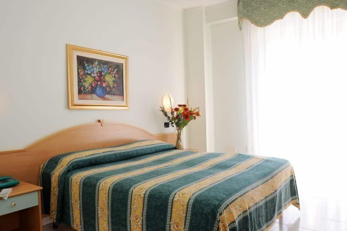 A hotel room with a bed, a nightstand, a framed painting, and a vase with flowers.