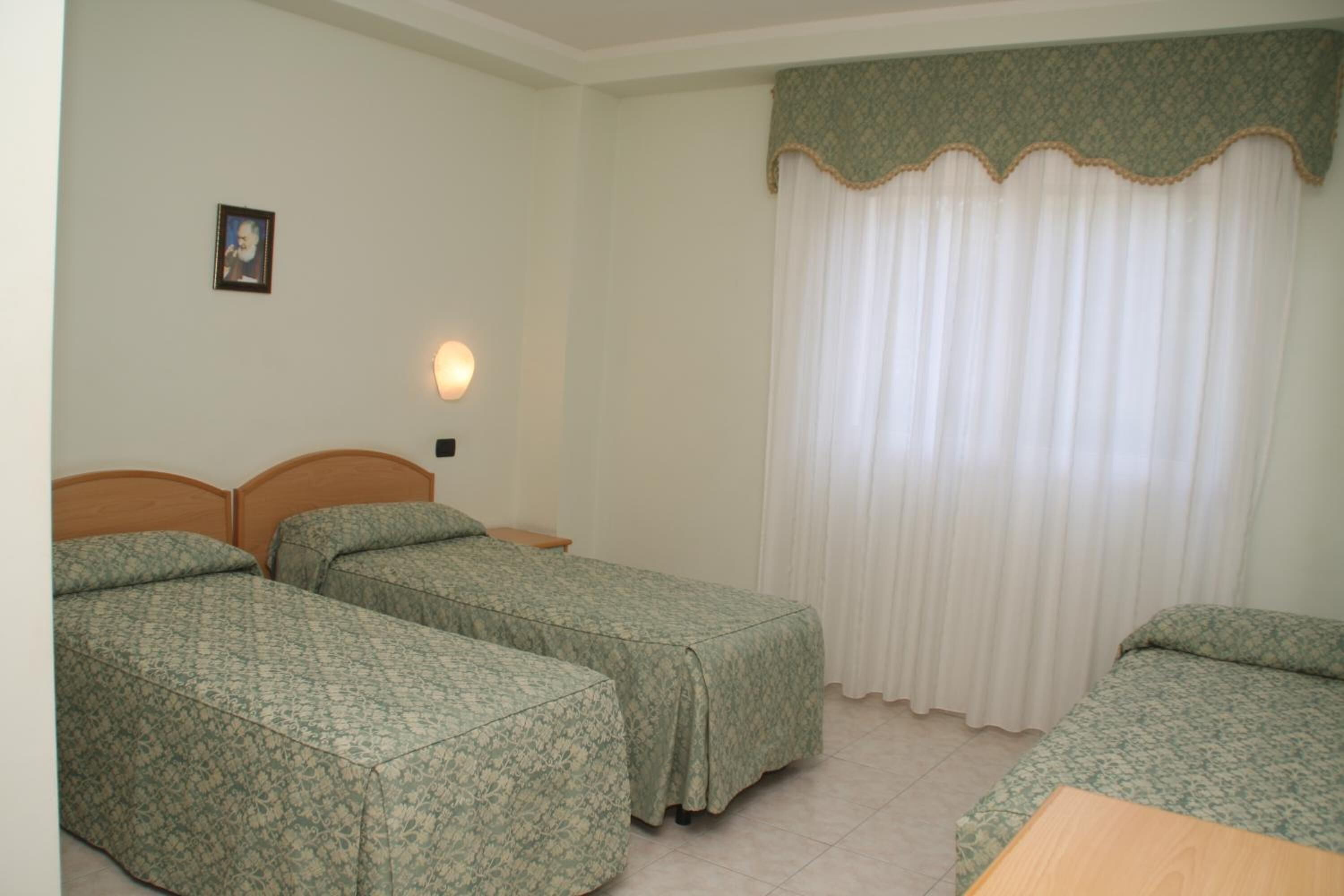 A room with two single beds, a wooden headboard, a nightstand, a wall-mounted lamp, and a large window with white curtains.