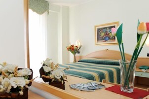 Una habitación de hotel con cama, una mesita de noche, un florero con flores, un cuadro en la pared y una ventana con cortinas.
