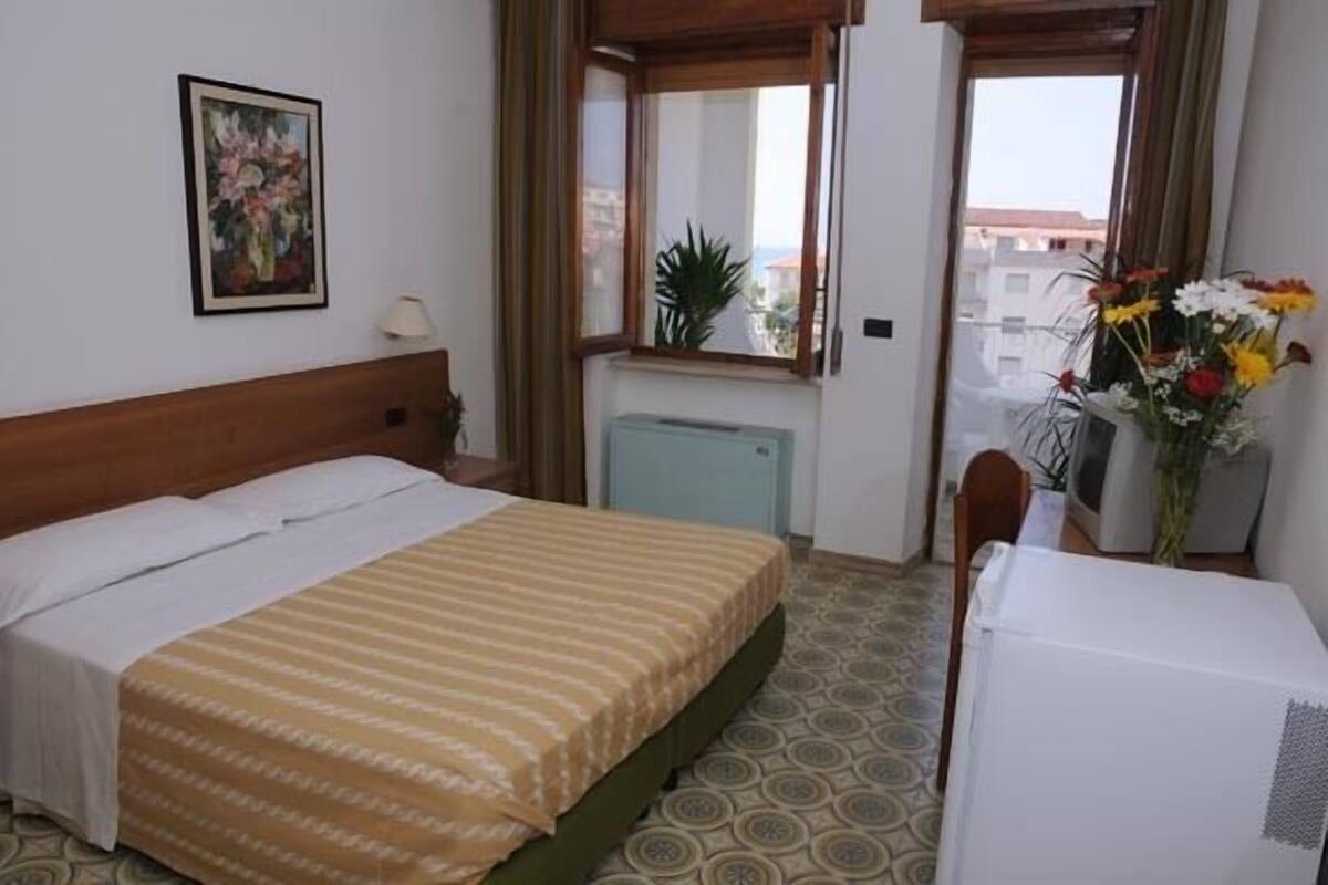 Habitación de hotel con cama, una mesita con un jarrón de flores, un radiador y una ventana con vistas a edificios.
