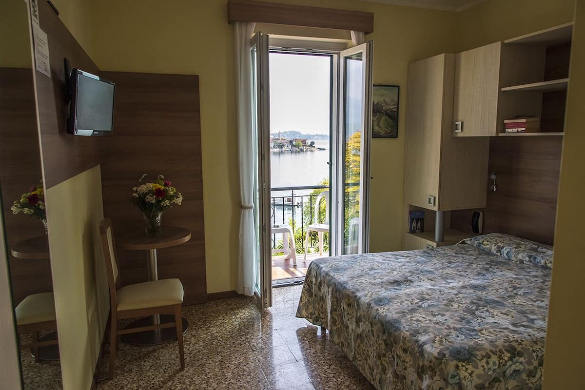 Une chambre avec un lit, un bureau et une chaise, une petite table avec des fleurs, une télévision et un balcon donnant sur l’eau.