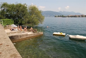 A lakeside swimming area with a concrete platform, a small boat, and a distant mountain range. - Hotel Villa Ruscello (Baveno)