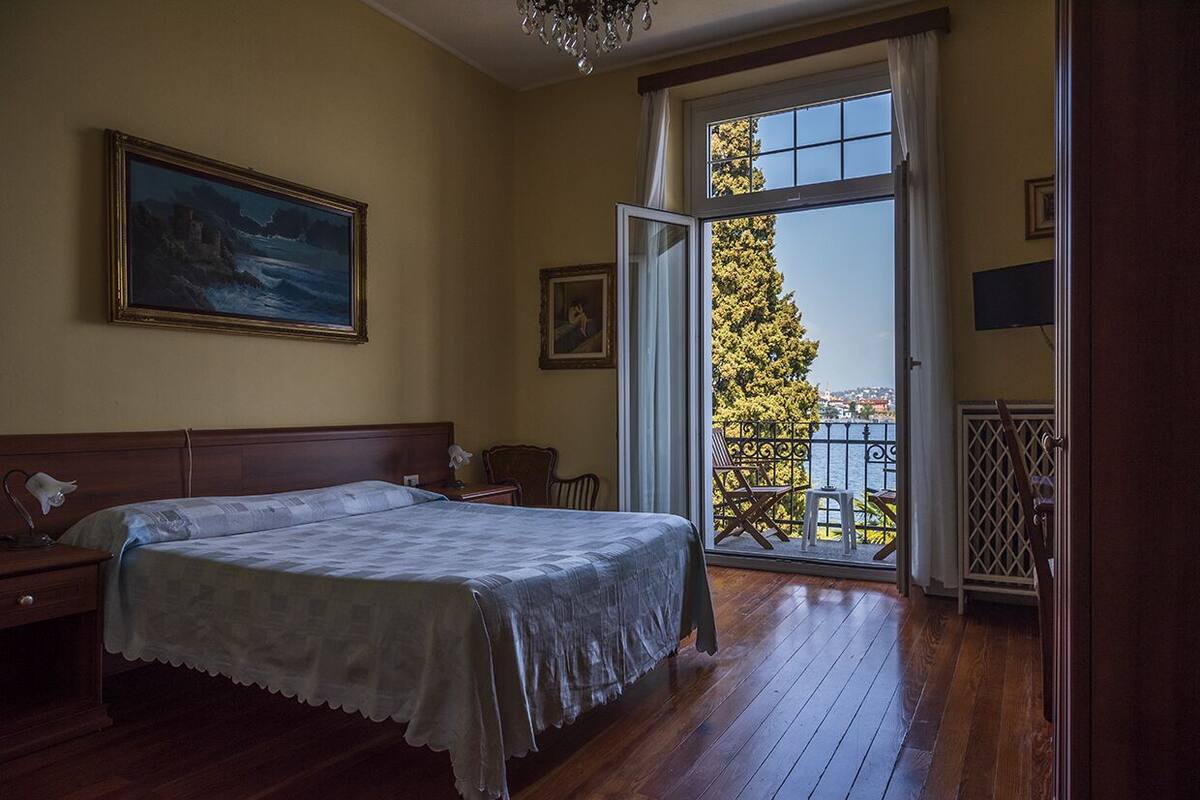Une chambre avec un grand lit, une table de chevet, un bureau, une chaise, un tableau et un balcon donnant sur la mer.
