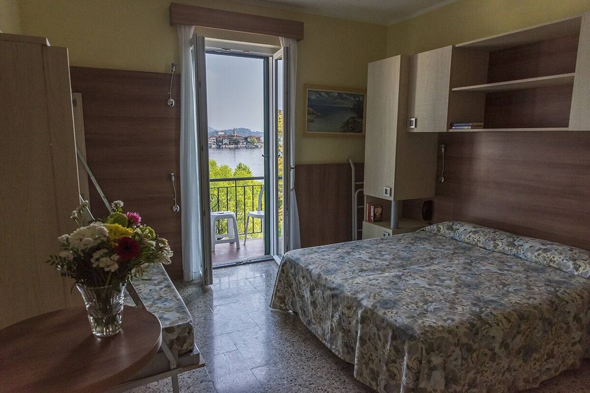 Une chambre à coucher avec un lit, un bureau, une chaise, un vase de fleurs et une vue sur une étendue d’eau, visible à travers une porte ouverte.