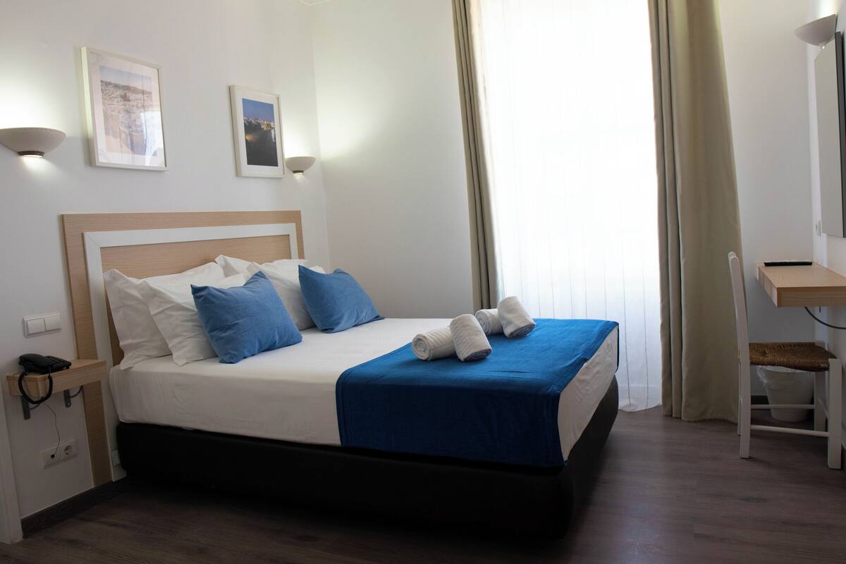 A neatly made bed with blue and white bedding, a bedside table with a lamp, and a desk with a chair.