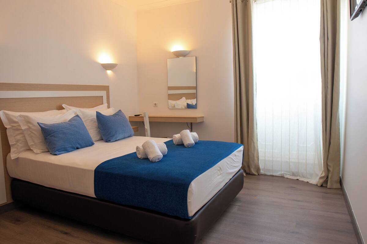A neatly made bed with a blue blanket, white pillows, and two rolled towels. There is a bedside table with a lamp and a mirror above it. The room has wooden flooring and beige curtains on the window.