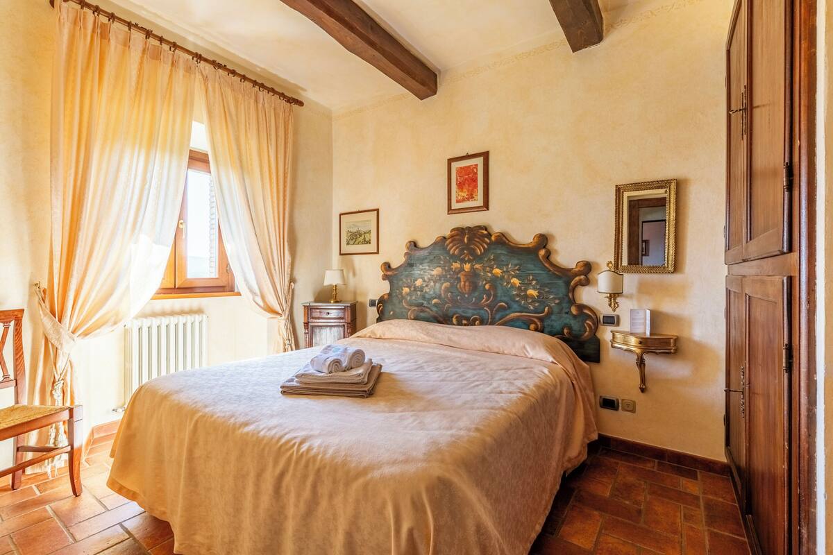 A bedroom with a large bed, a wooden headboard, a nightstand, a lamp, a window with curtains, and a radiator.