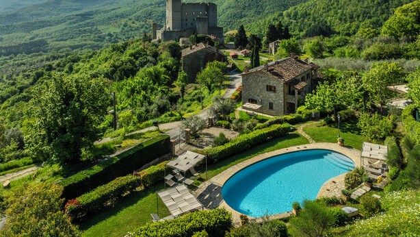 Un castillo rodeado de exuberante vegetación, una piscina y un edificio de piedra.