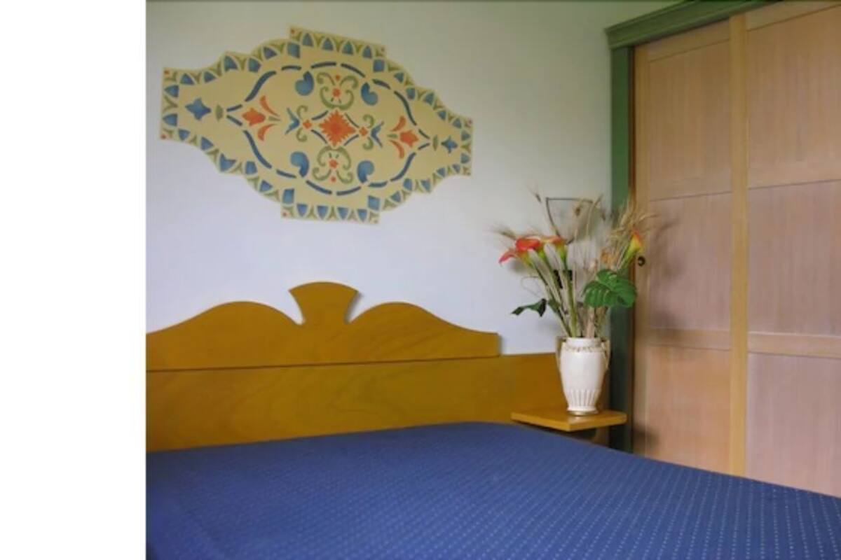 A bedroom with a wooden bed, a blue mattress, a decorative wall hanging, and a wooden wardrobe.